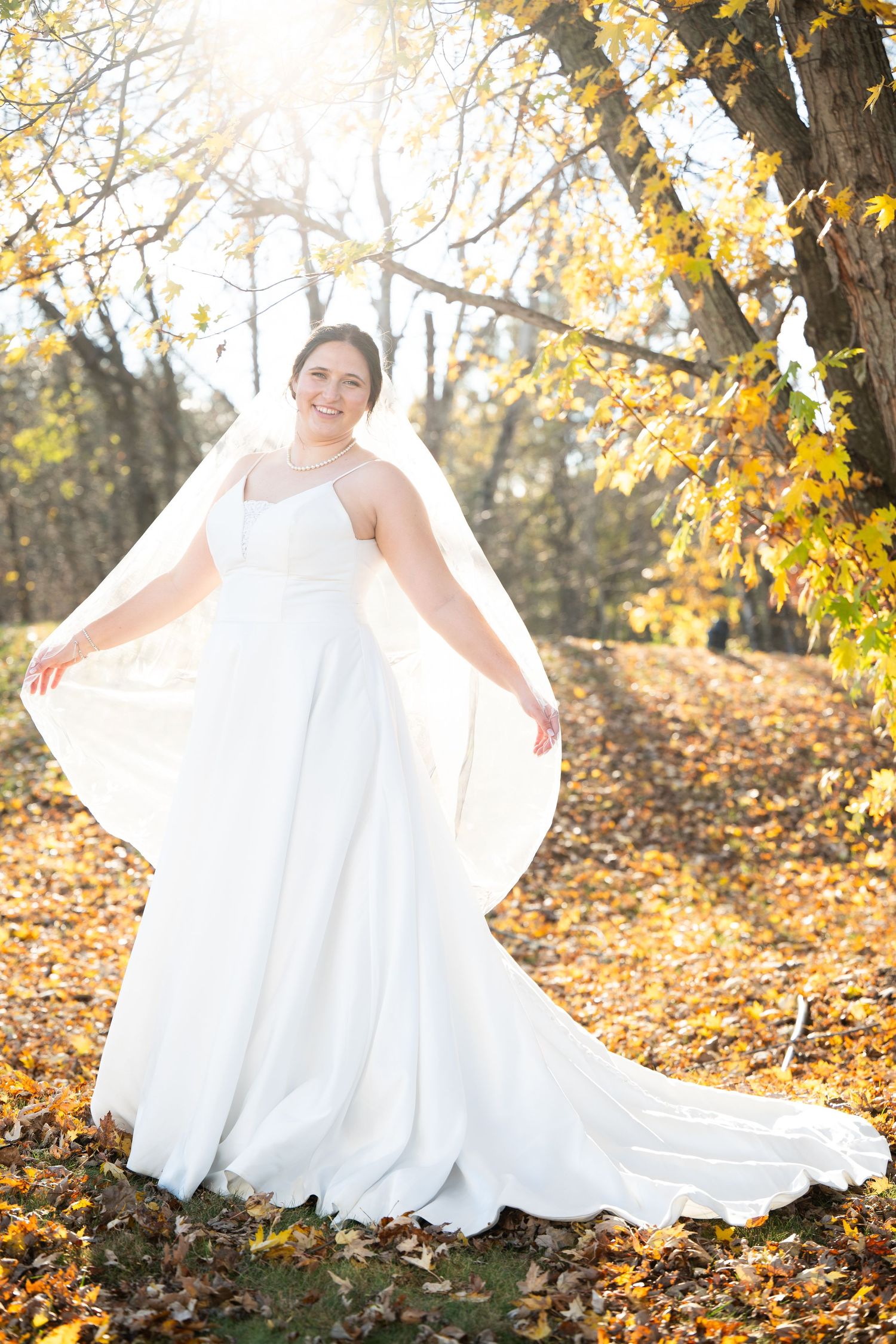 Bride in flowing white dress twirls among autumn leaves in golden sunlight.