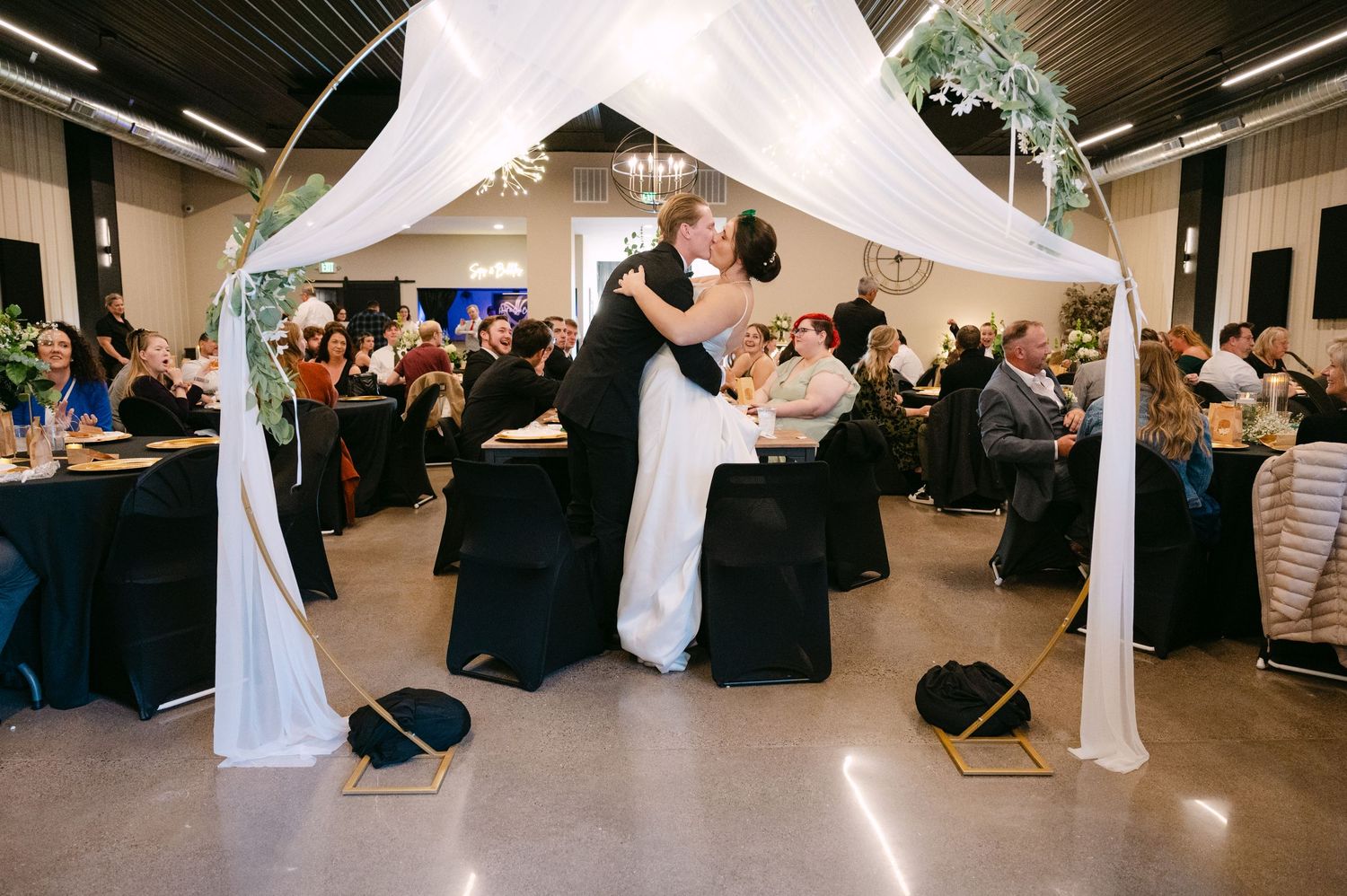 Large indoor wedding reception space with white fabric decor and black-covered chairs arranged around tables.
