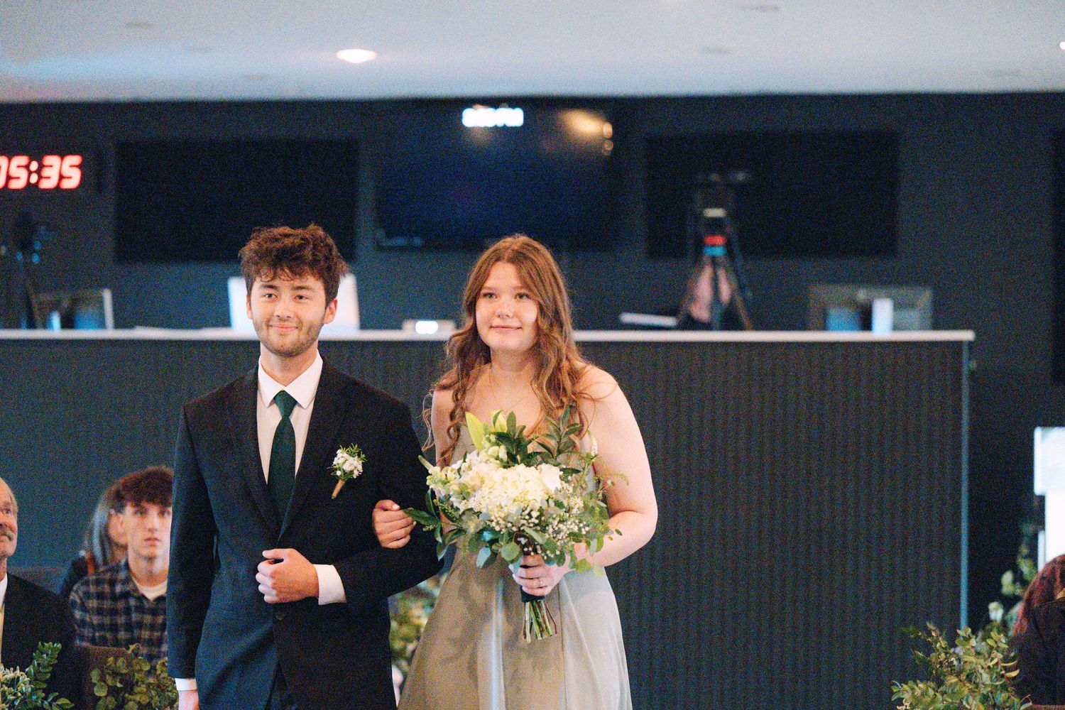 A couple walks down the aisle at a modern indoor wedding ceremony with the bride holding a white floral bouquet.