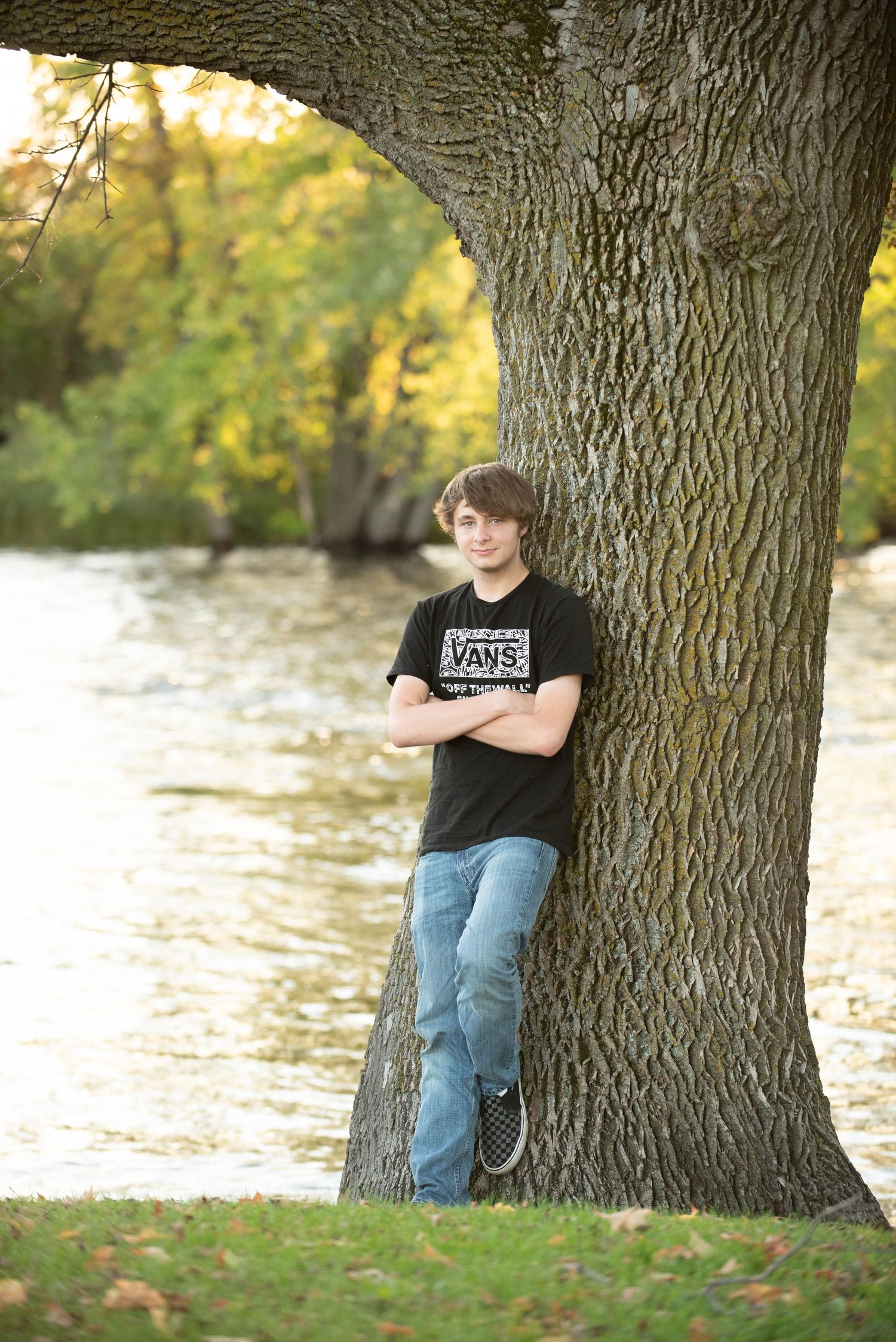 Casual portrait session against large tree trunk with water and autumn foliage in background.