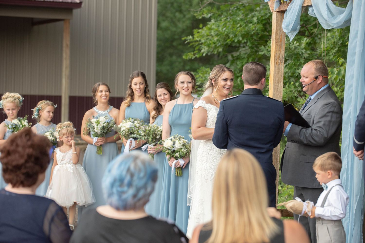 Wedding party stands at wooden arch decorated with light blue fabric during outdoor ceremony. Zimmerman MN Weddings