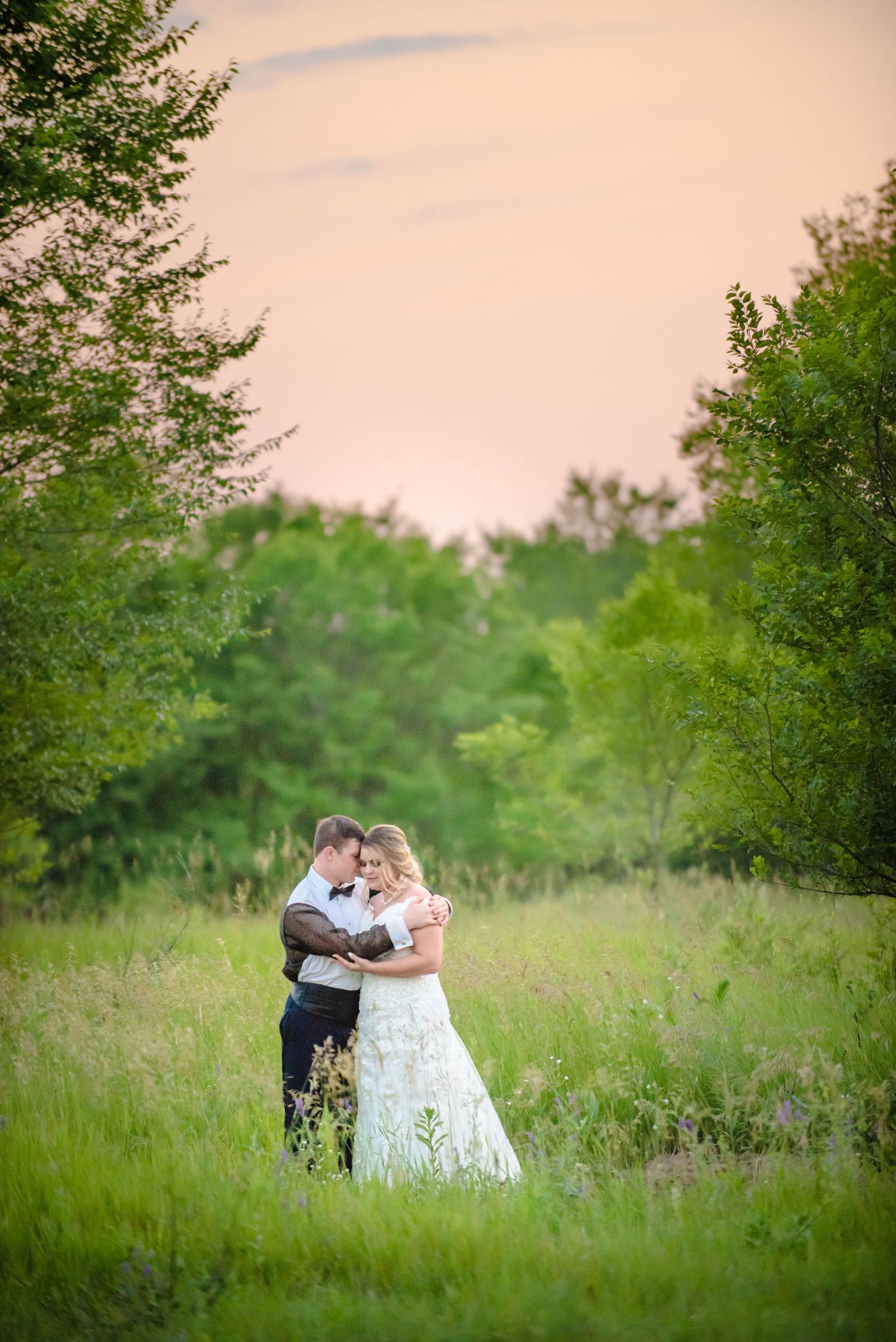 Romantic sunset portrait of newlyweds embracing in a meadow with green trees. Zimmerman MN Weddings