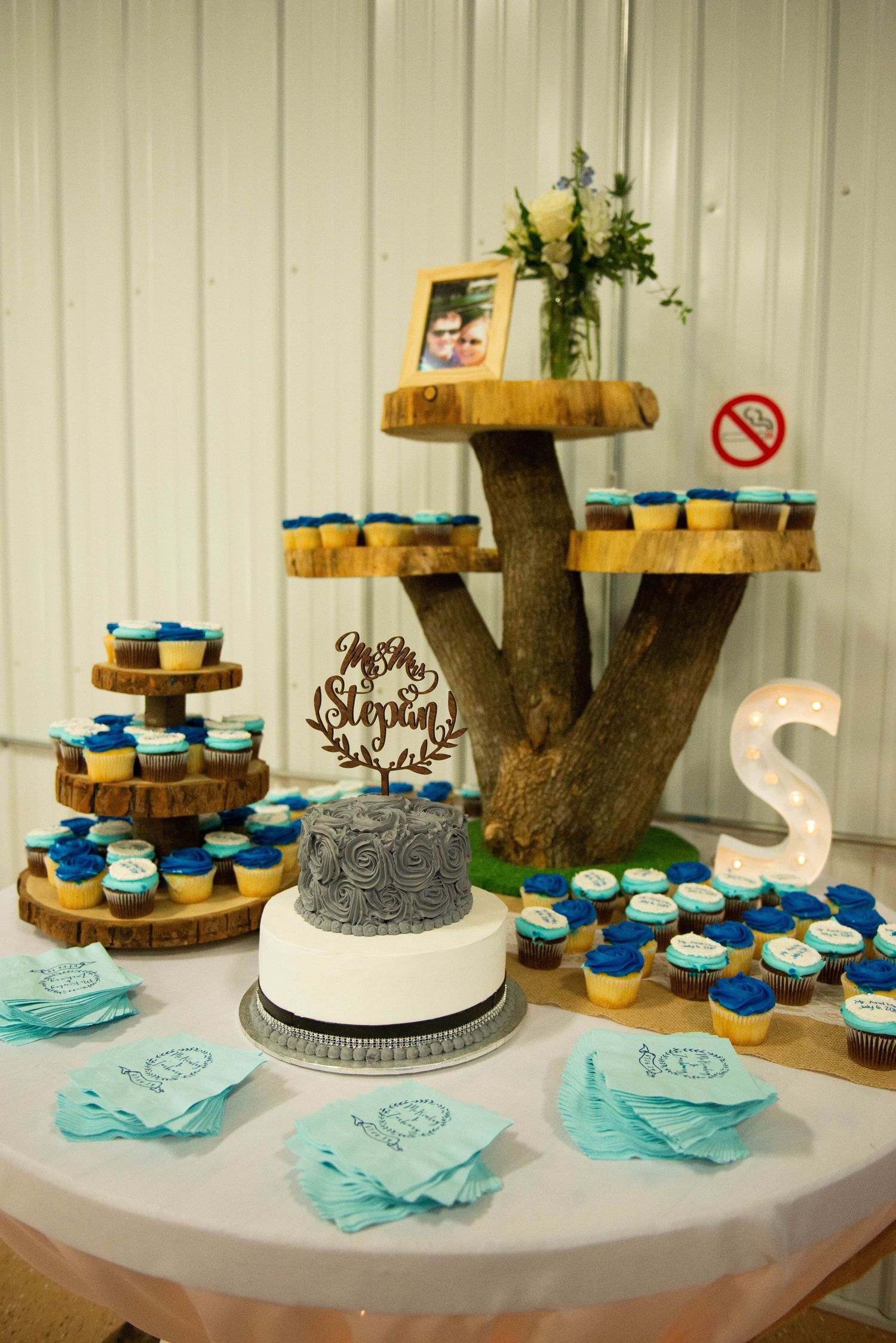 Rustic wedding dessert display featuring a tree trunk cupcake stand with blue frosted cupcakes and a gray and white wedding cake. Zimmerman