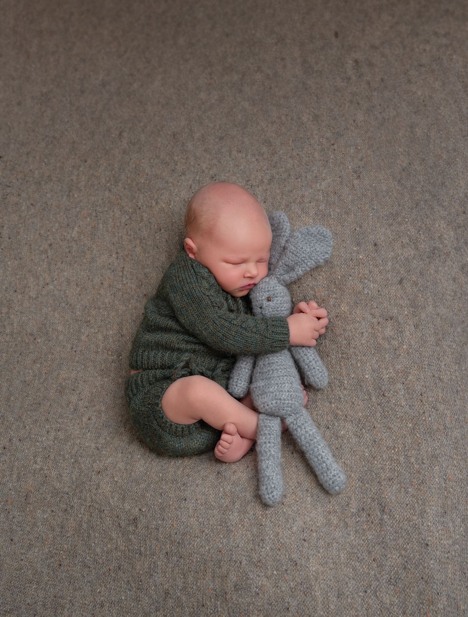 Minnesota Best newborn photographer - Baby in green knit sweater cuddles with gray stuffed bunny toy while lying on carpet.