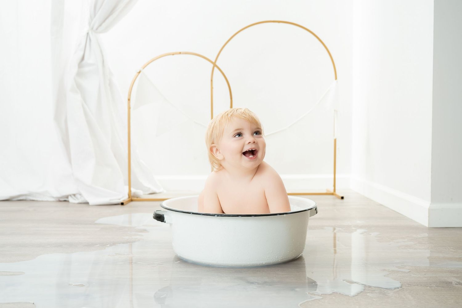 Happy baby sits in vintage white enamel basin with gold arched backdrop against a bright airy background. Princeton MN Milestone, Maternity