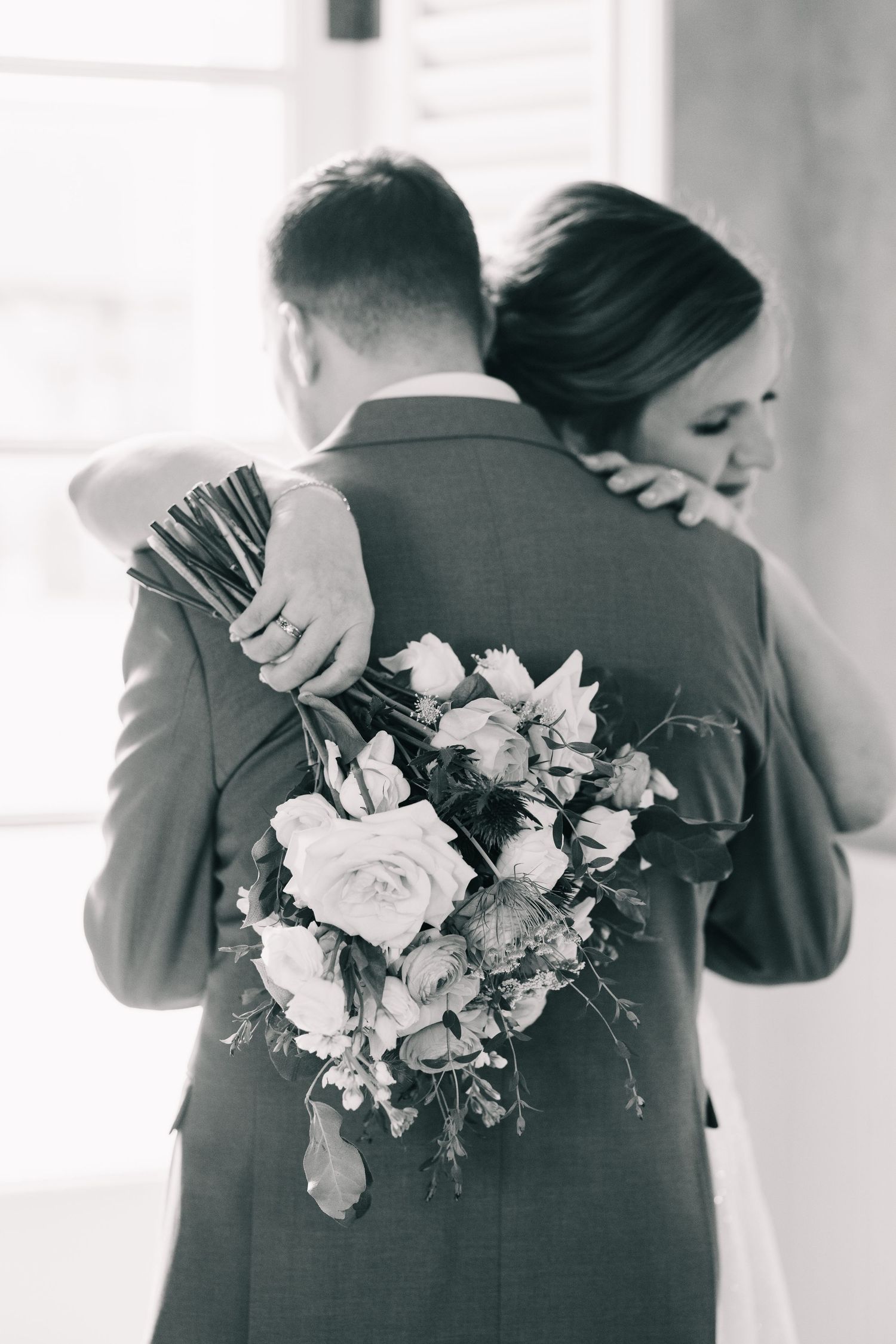 Black and white photo of bride and groom embracing, bride holding elegant floral bouquet with roses.