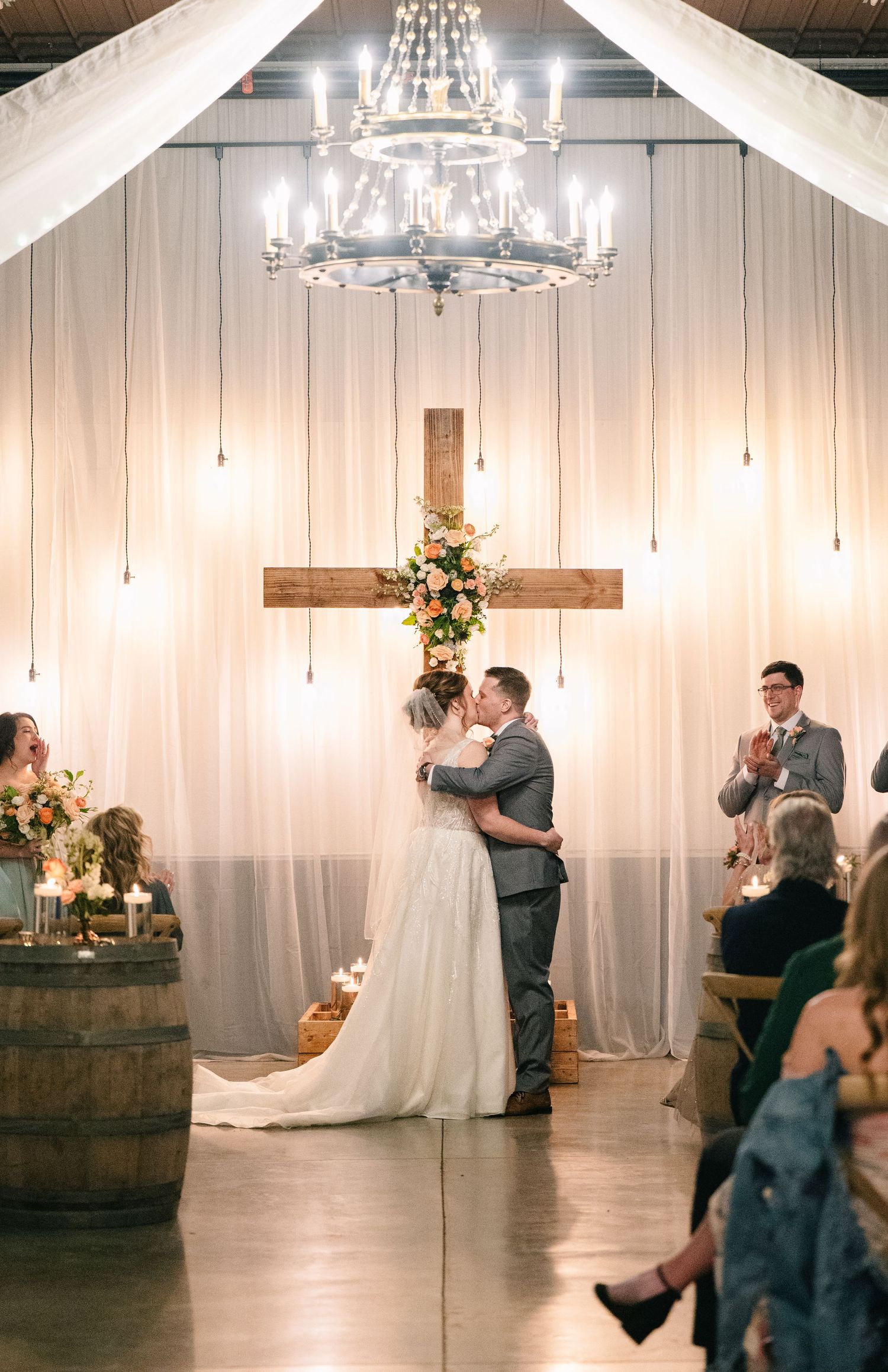 Bride and groom share first kiss at rustic wedding ceremony with wooden cross, chandelier, and white draping backdrop.