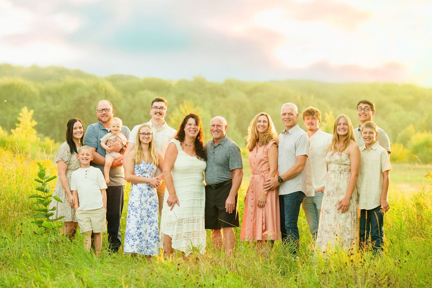 Large family group portrait outdoors in a sunny meadow with lush green trees in the background.