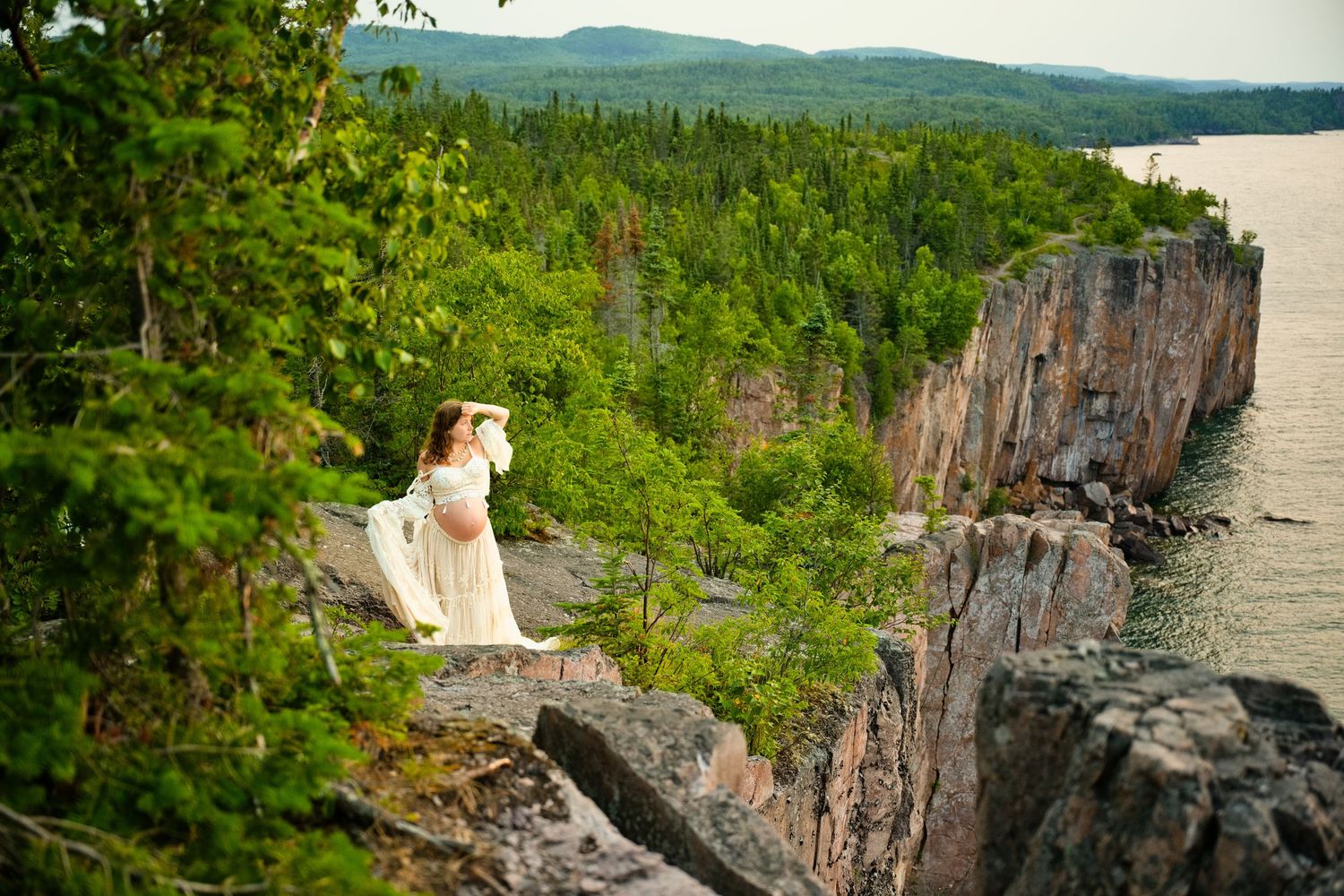 Bride in white gown standing on rocky cliff overlooking lush forest and serene lake in scenic outdoor wedding photo.