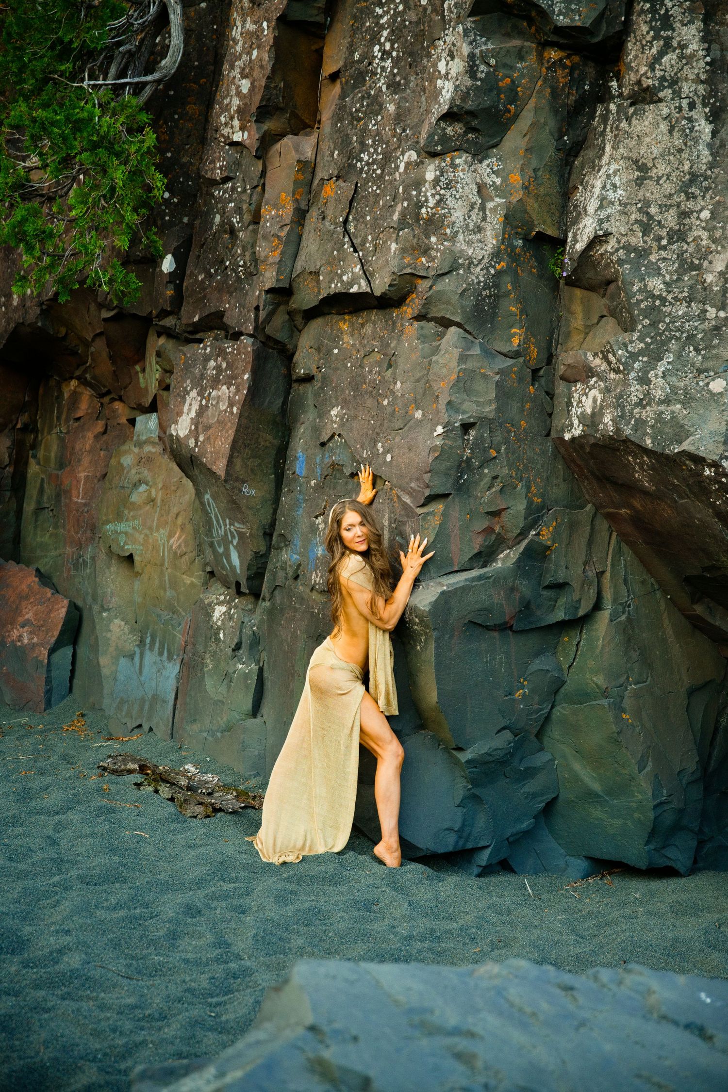 Woman in golden dress poses gracefully against dramatic rocky cliff face on dark sandy beach.