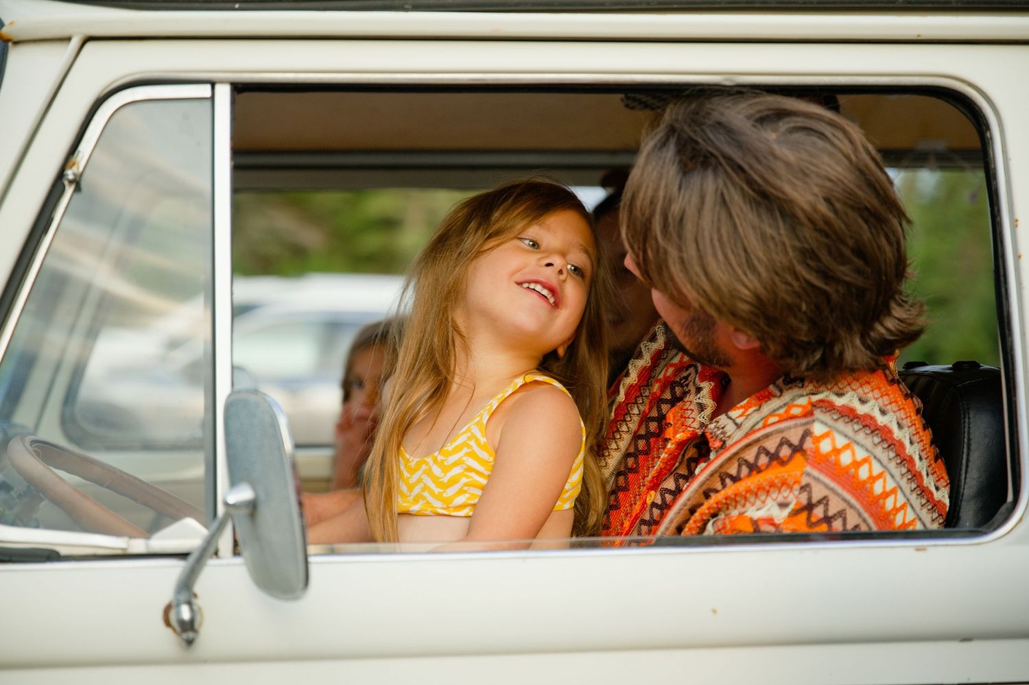 A young girl smiles at an adult in a vintage van, both wearing colorful clothing on a road trip.