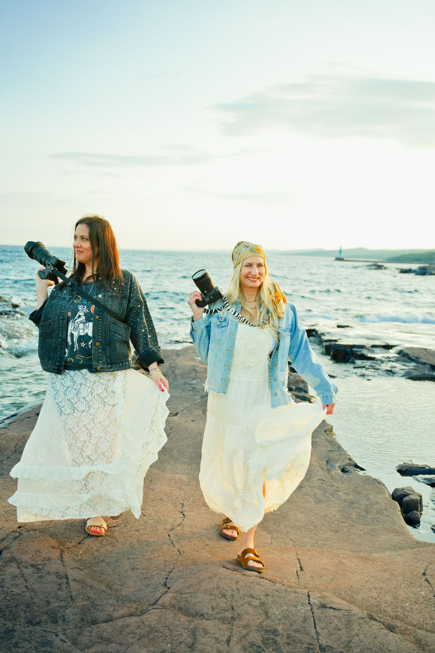 Two women in white dresses and denim jackets hold cameras on rocky coastal shore at sunset.