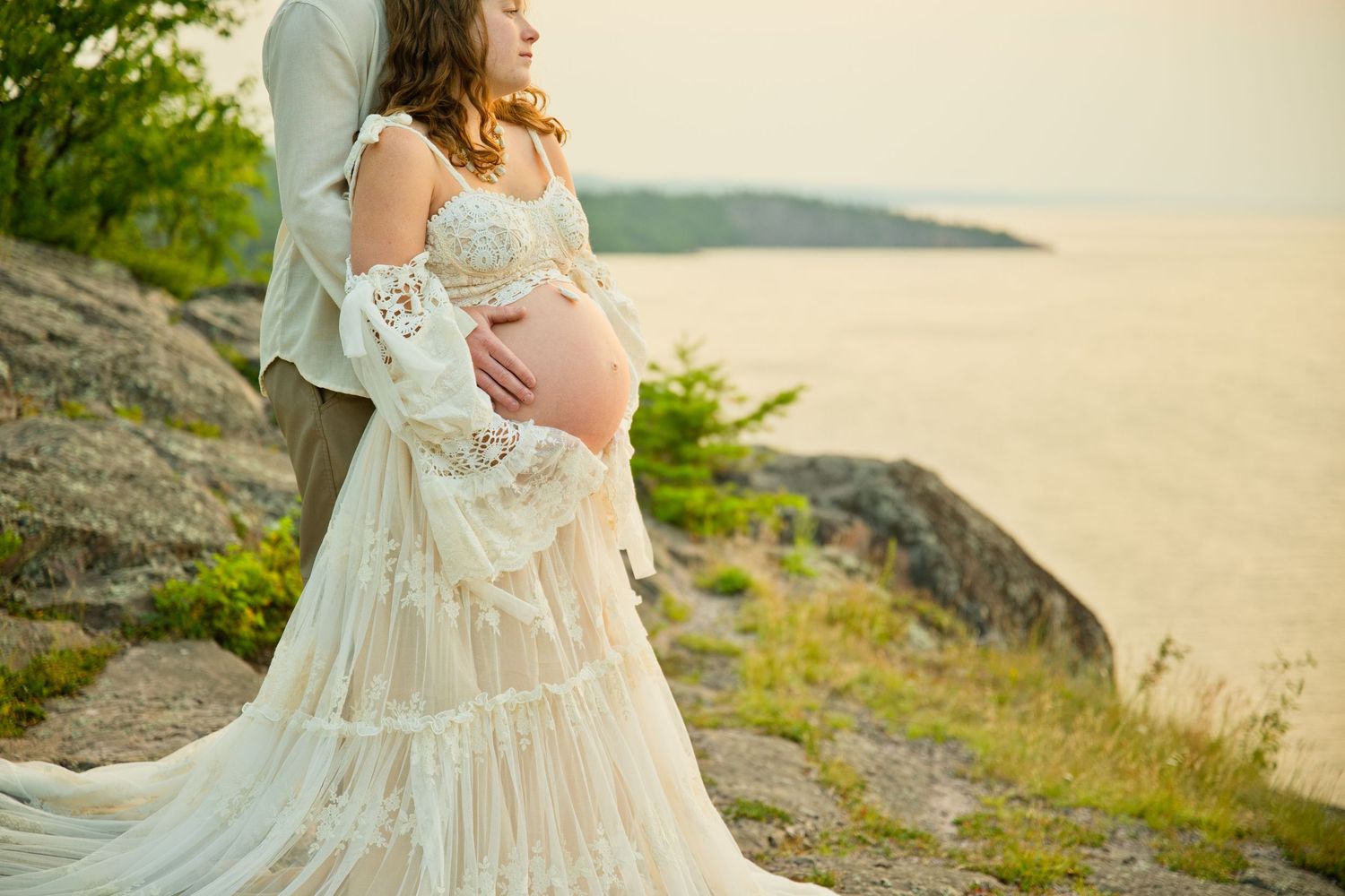 Pregnant woman in white lace gown embraced by partner on rocky overlook with scenic water view.