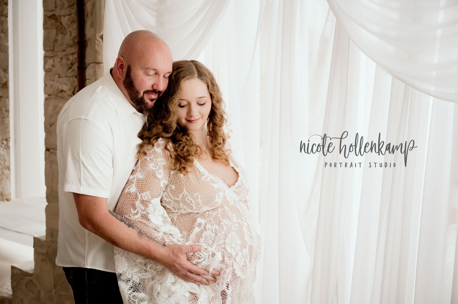 Intimate maternity portrait of couple embracing against white curtain backdrop.