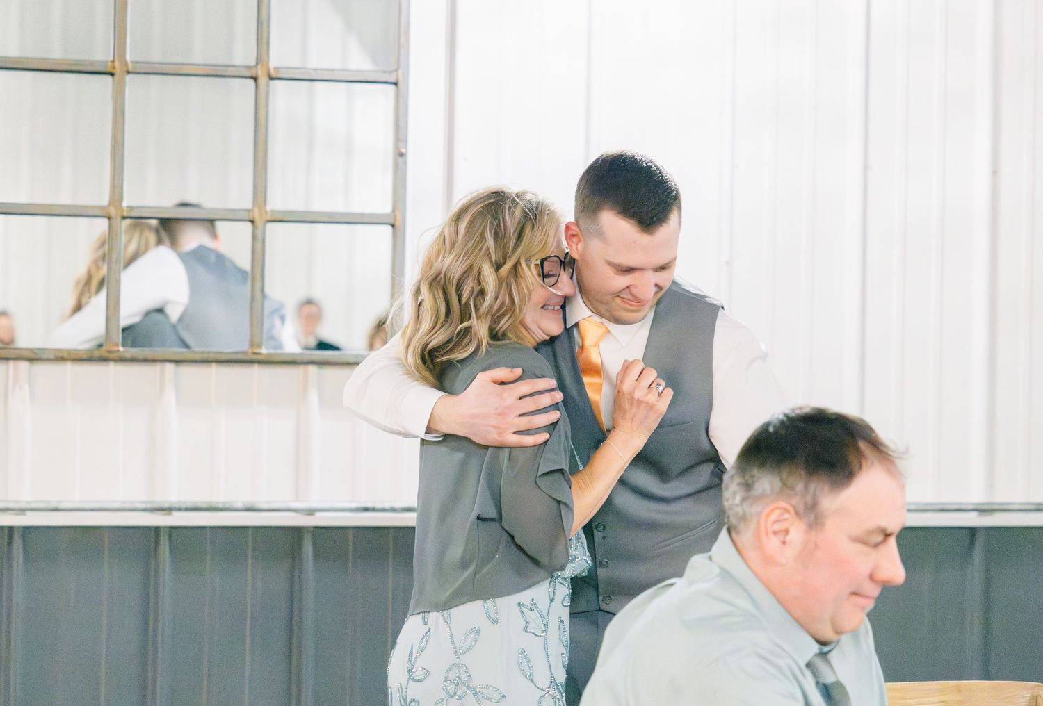 Woman hugs groom in gray vest at wedding reception while another guest sits in foreground.