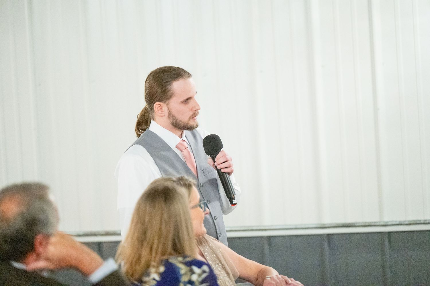 Groomsman with long hair in gray vest delivers wedding speech into microphone at reception.