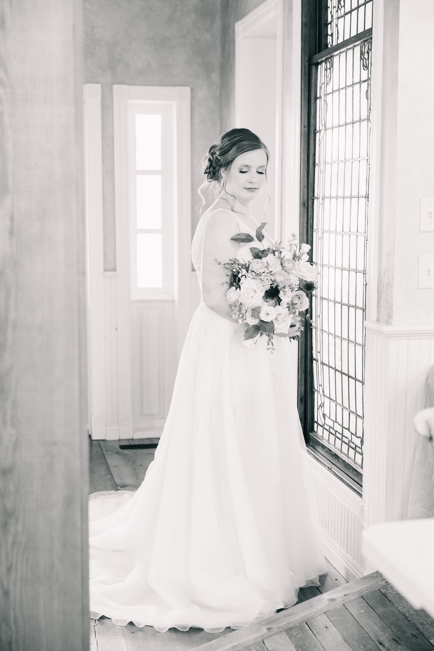 Black and white photo of bride in elegant backless gown holding bouquet beside latticed window.