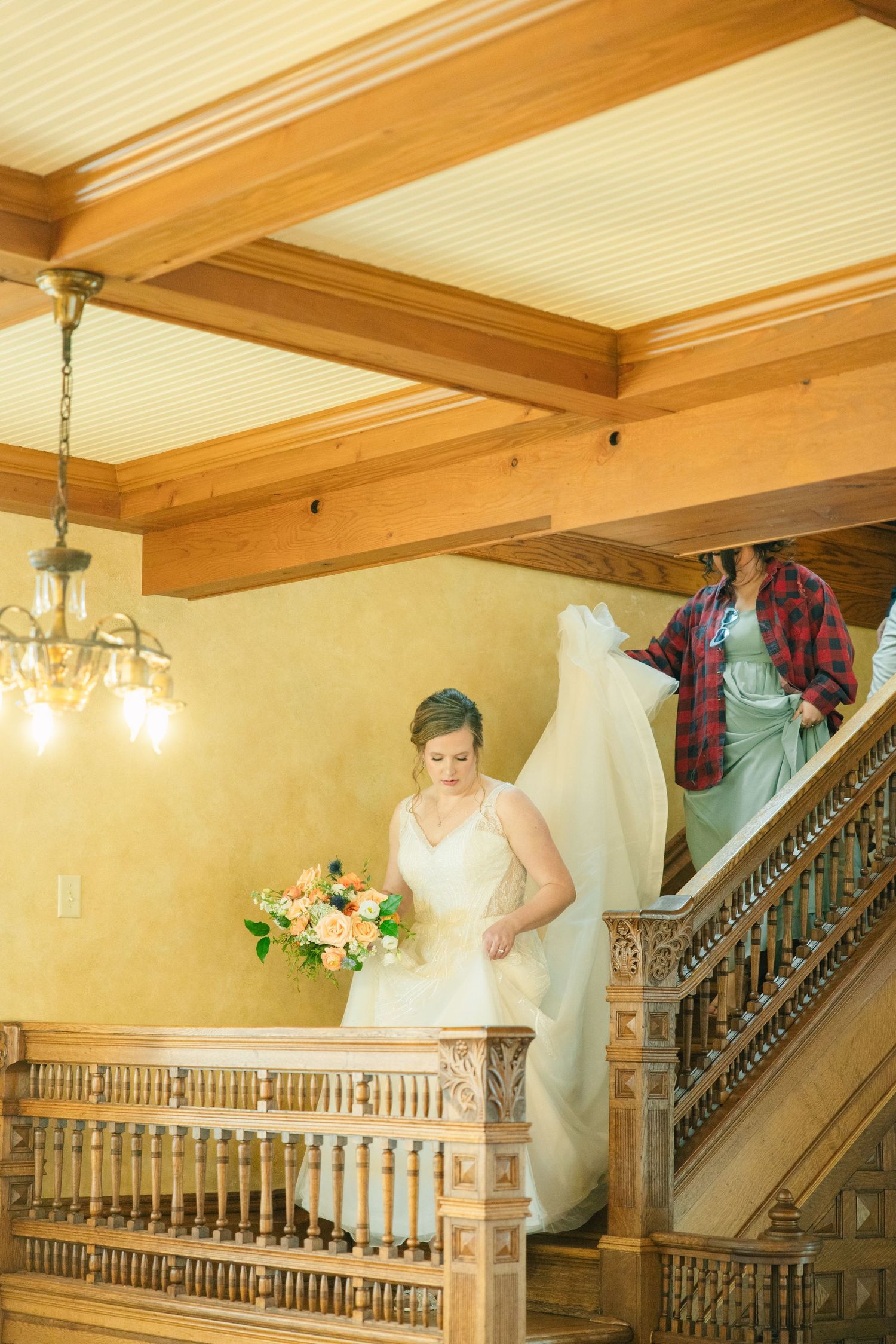 Bride descends ornate wooden staircase as assistant holds her flowing white gown train.