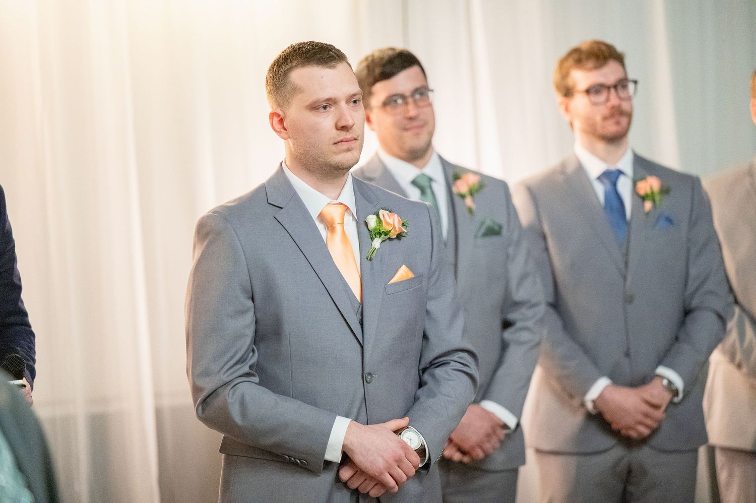 Groom and groomsmen stand at altar in matching gray suits with orange boutonnieres during wedding ceremony.