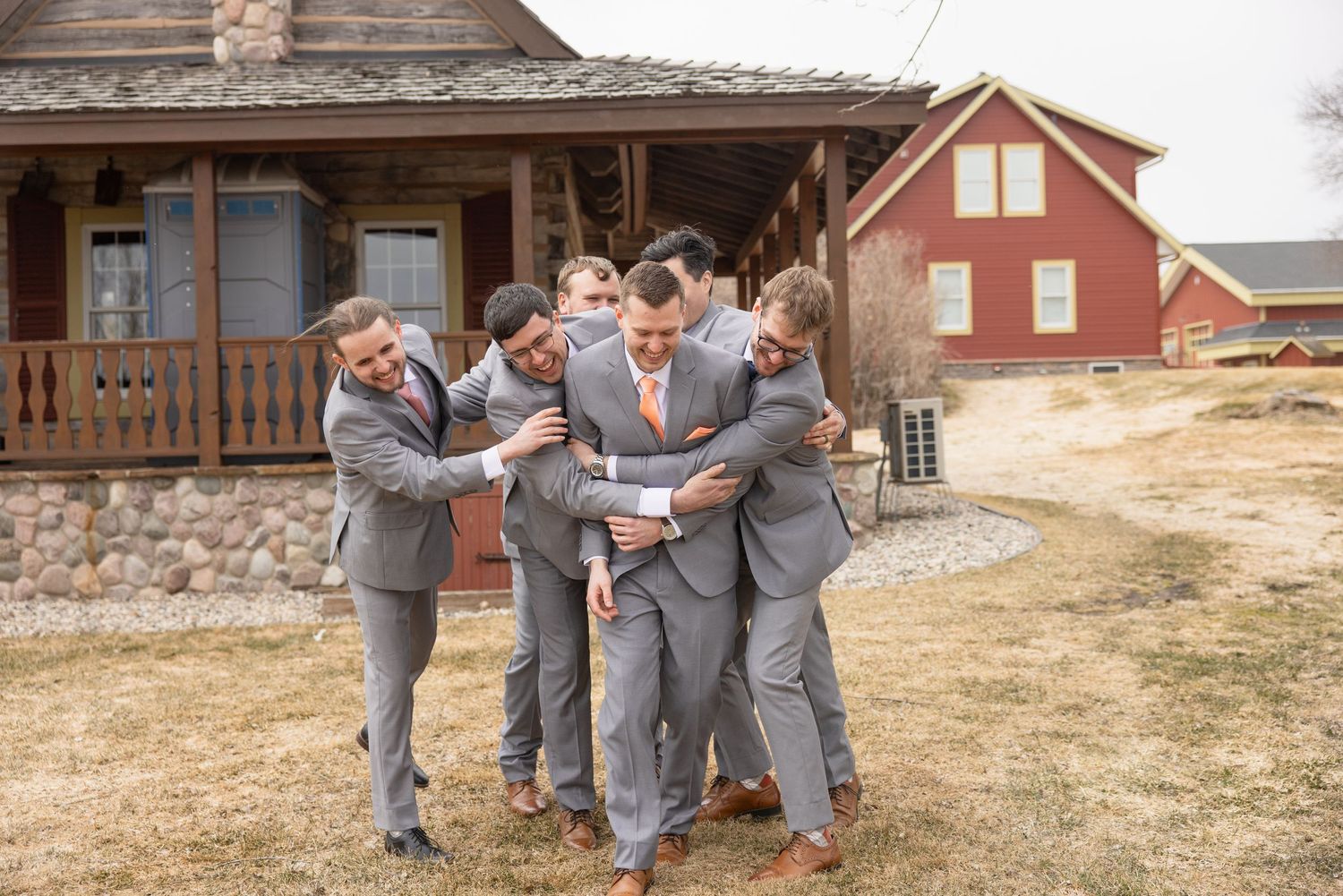 Groomsmen in gray suits with peach ties laughing together outside a rustic wedding venue.