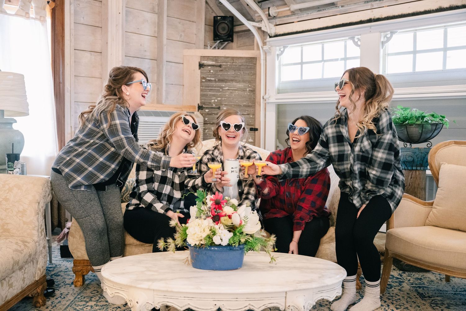 Five bridesmaids in plaid shirts toast with champagne glasses while laughing together in a rustic venue.
