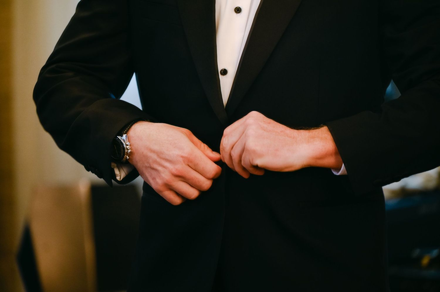 Groom adjusts his bowtie while wearing a classic black tuxedo in an elegant indoor setting.