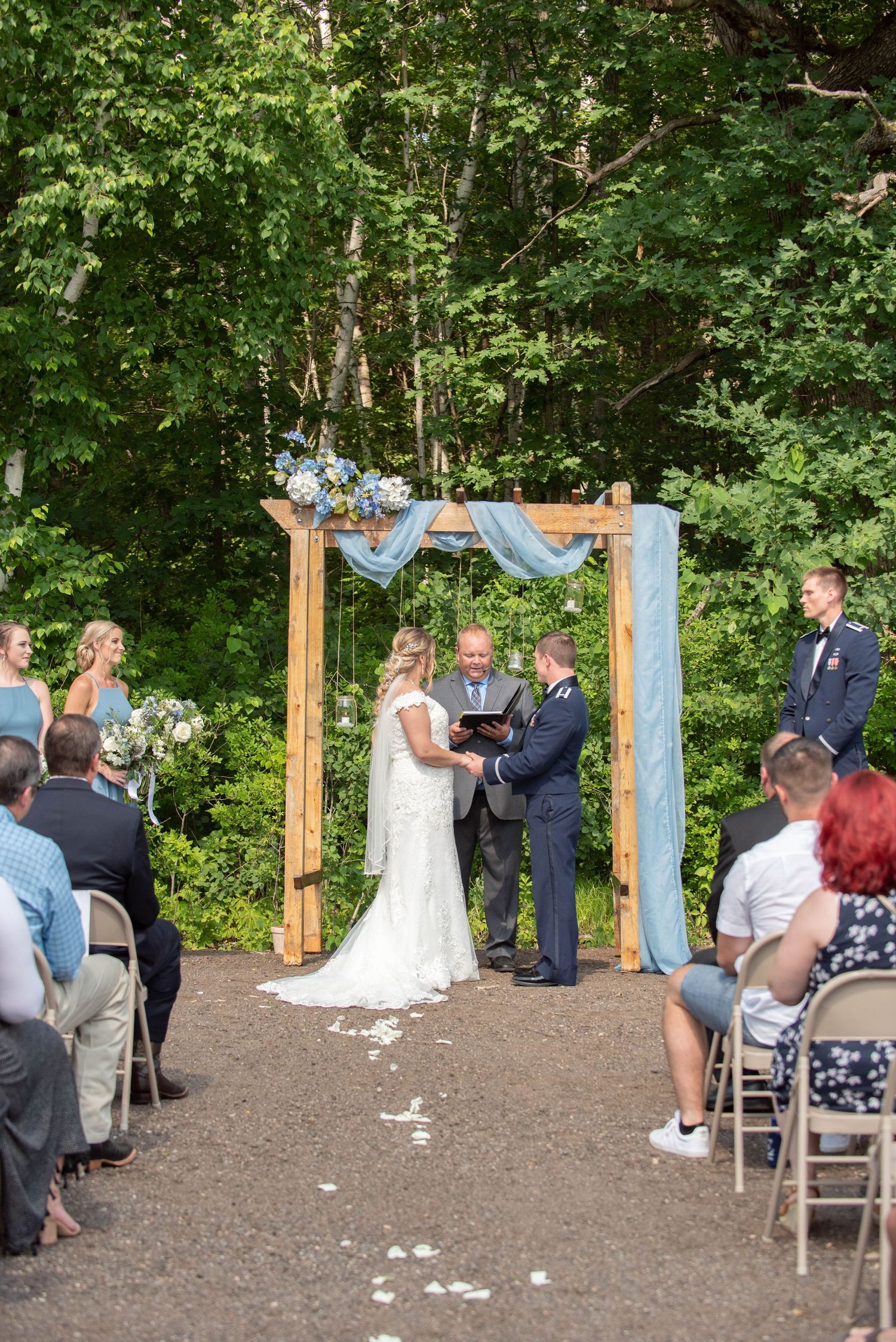 An outdoor wedding ceremony taking place under a wooden arch draped with light blue fabric in a wooded setting. Zimmerman MN Weddings