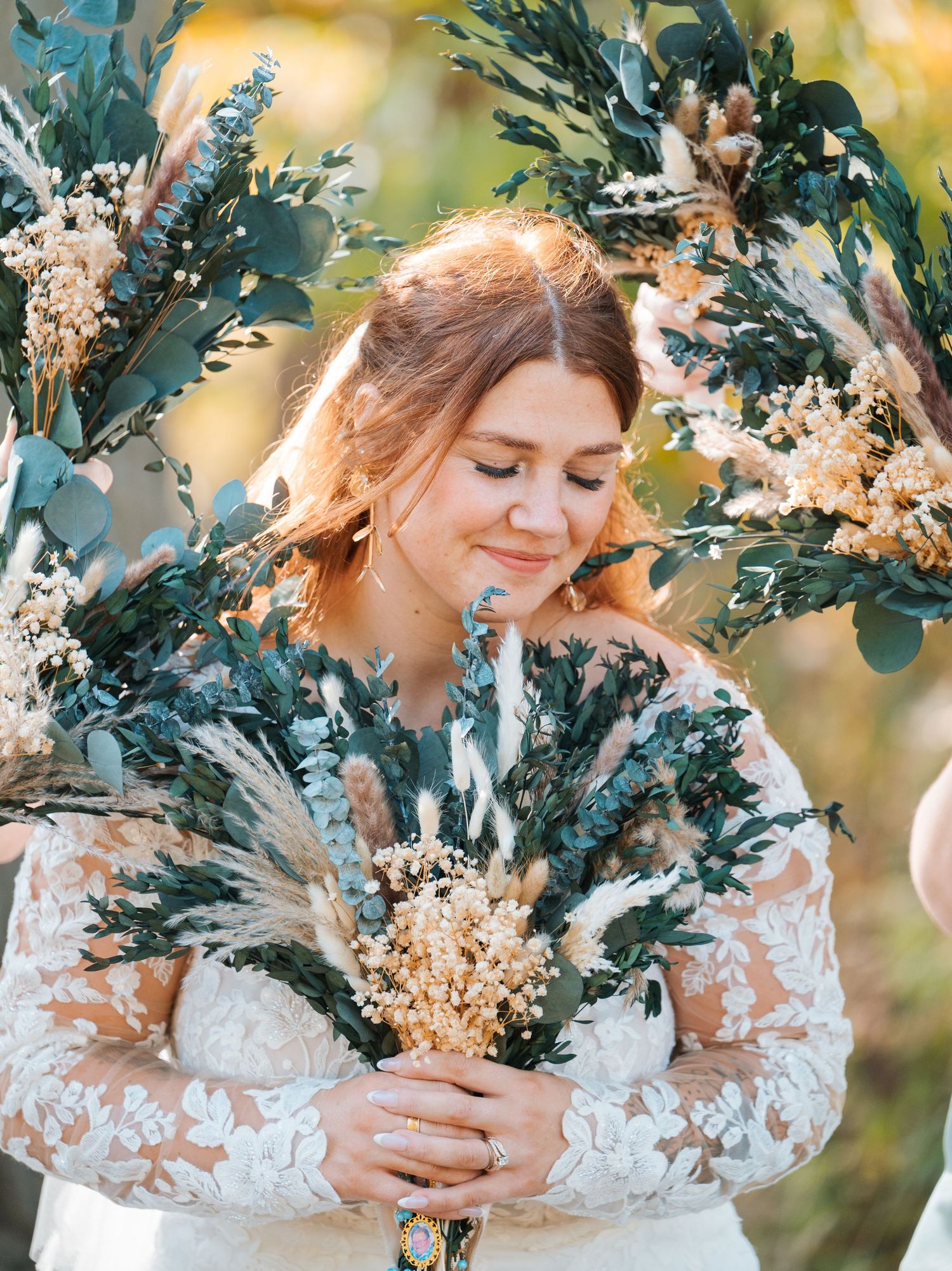 Bride in lace wedding gown smiles softly while holding a dried botanical bouquet with eucalyptus.