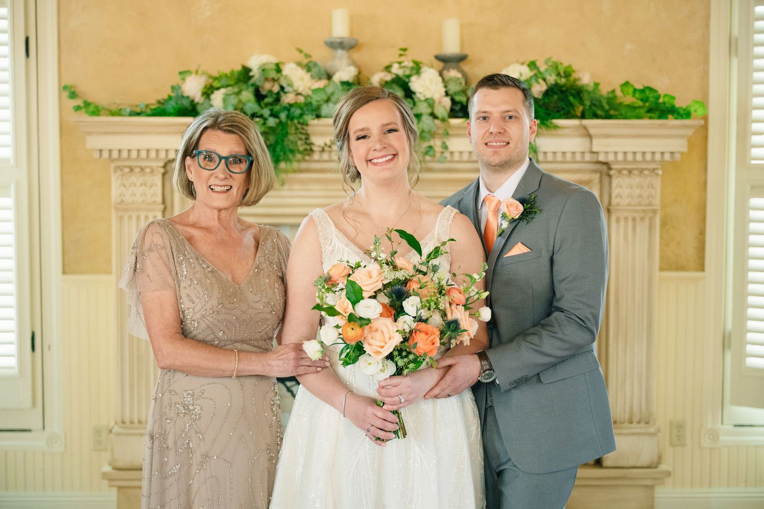 Bride with peach bouquet poses with groom in gray suit and older woman before floral fireplace.