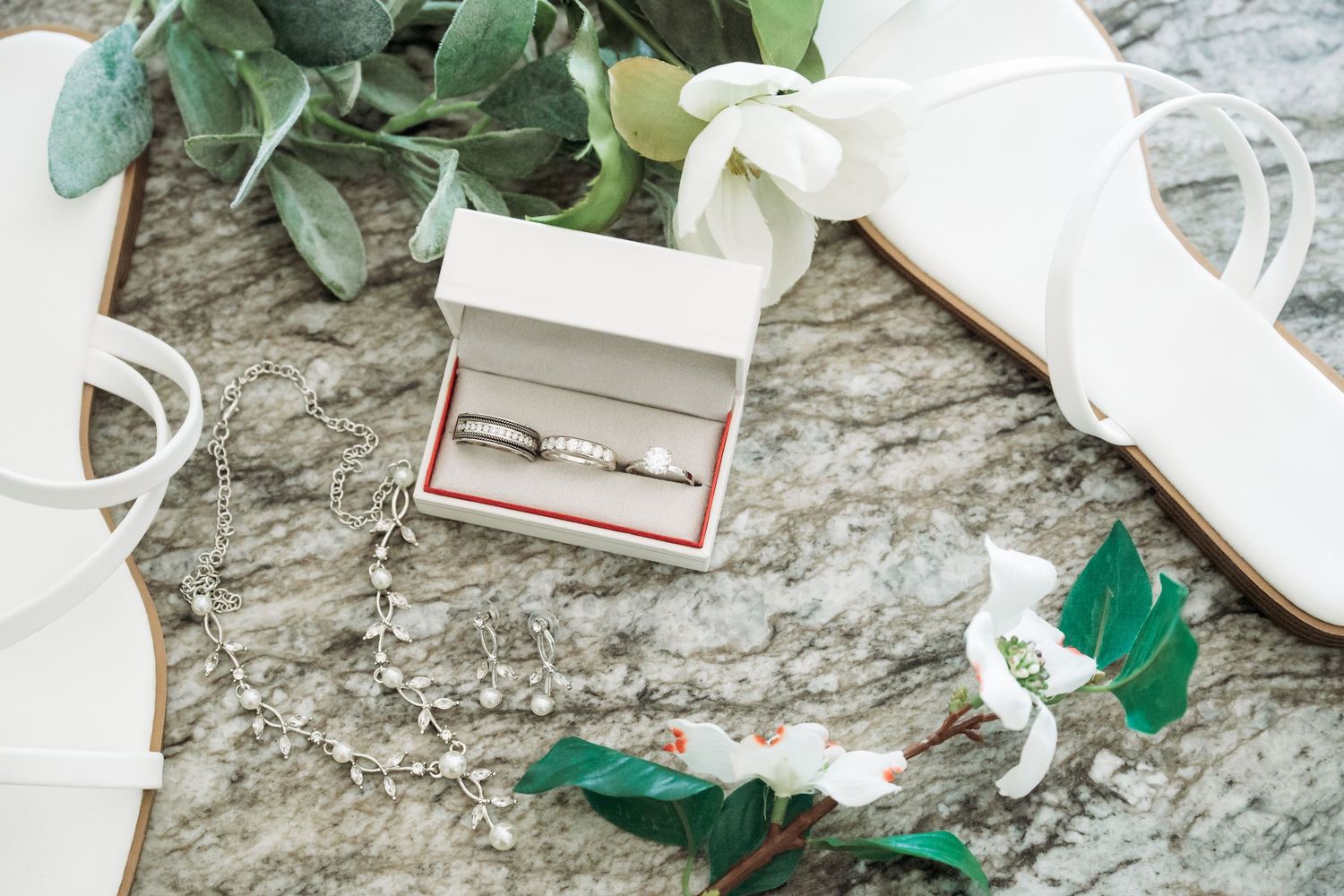Wedding rings in a white box surrounded by bridal accessories, white heels, greenery, and a delicate necklace on marble.