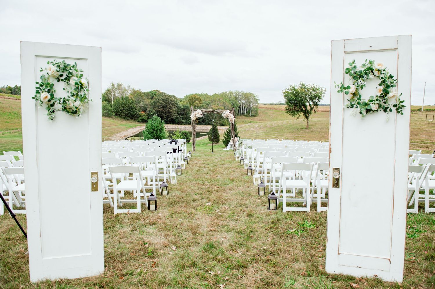 White vintage doors and chairs line grassy outdoor wedding ceremony aisle with floral decorations.