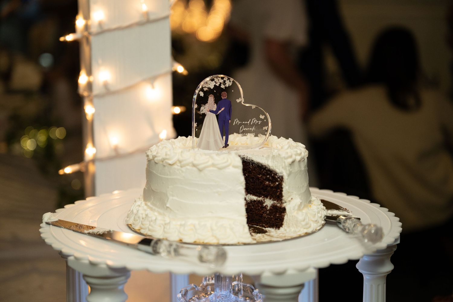 A white wedding cake with chocolate layers and a decorative heart topper displayed on an elegant stand.