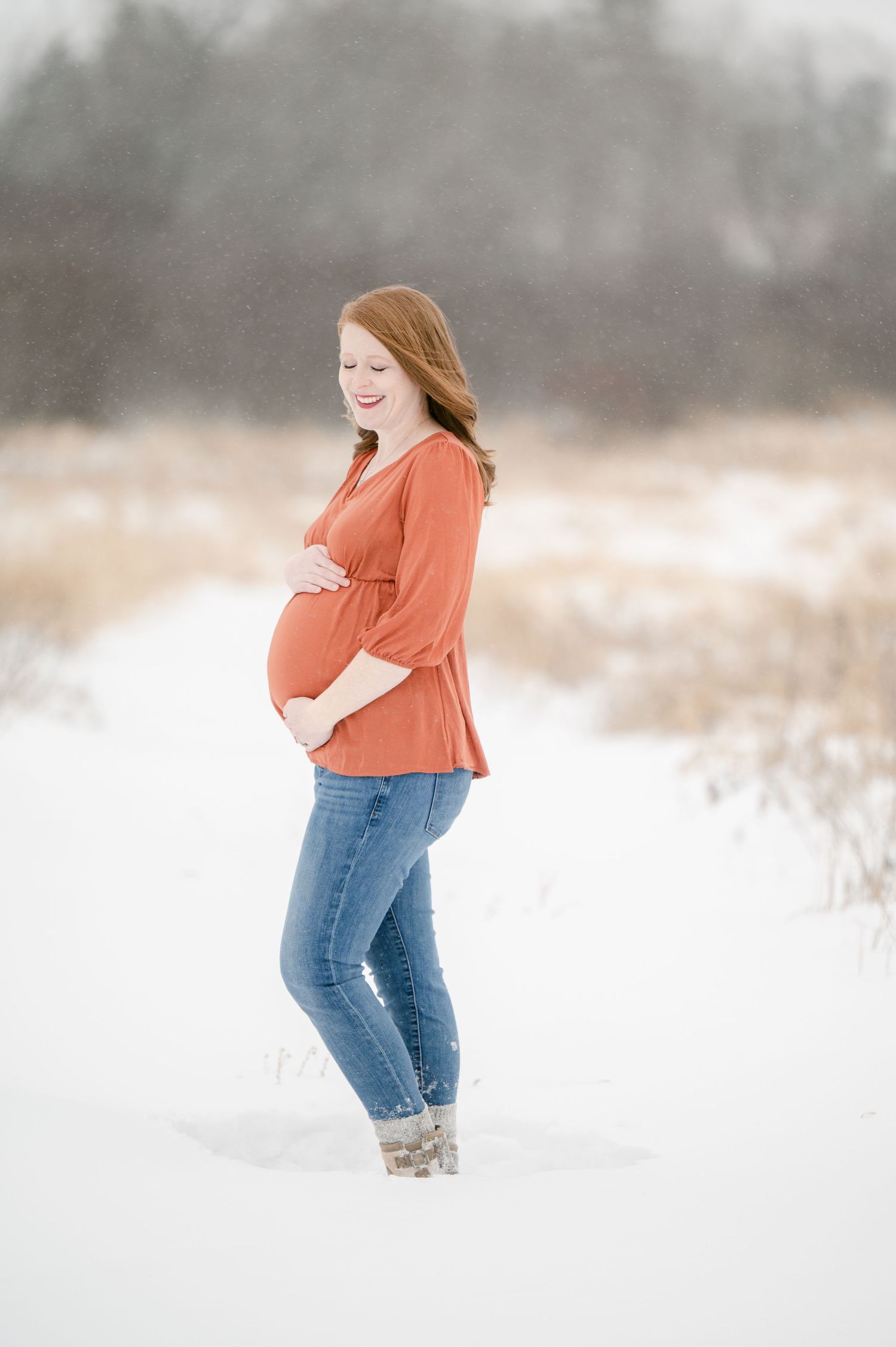 The Clark’s romantic winter maternity photoshoot in an orange top and jeans walking playfully through a snow-covered winter landscape.