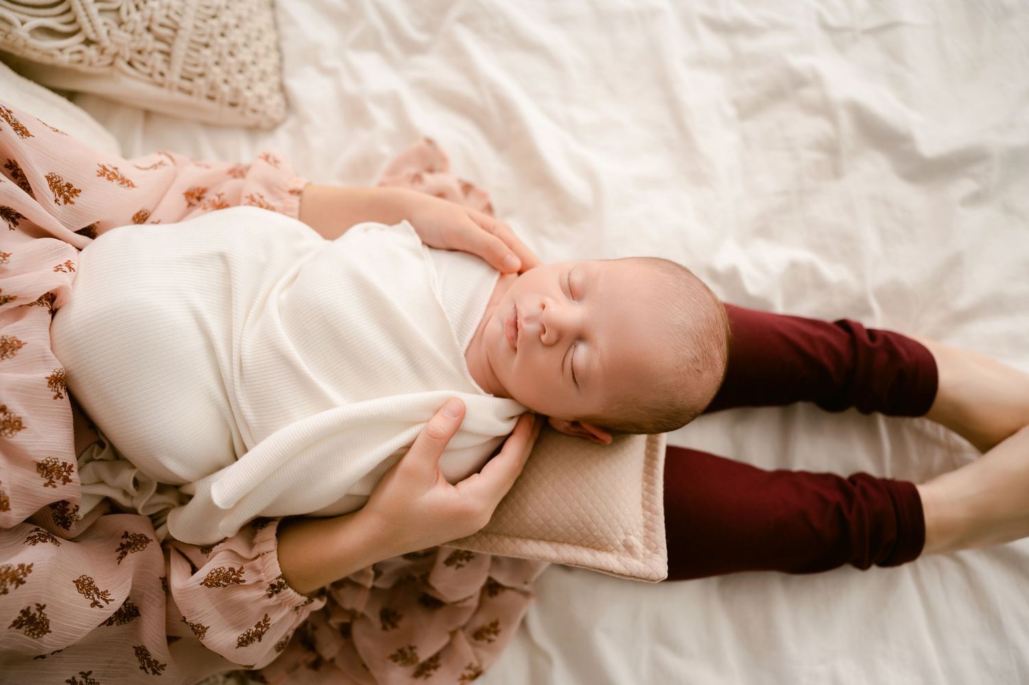 Sleeping infant wearing white onesie and burgundy socks rests peacefully on soft floral bedding.