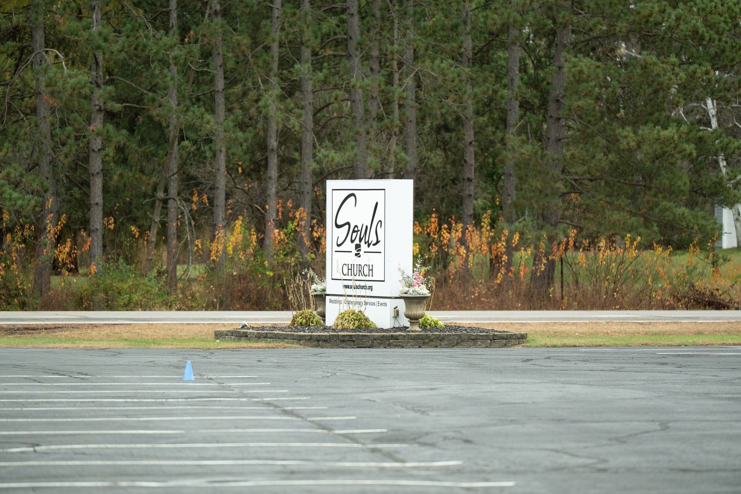 Small white memorial sign surrounded by orange flowers and trees along an empty parking lot.