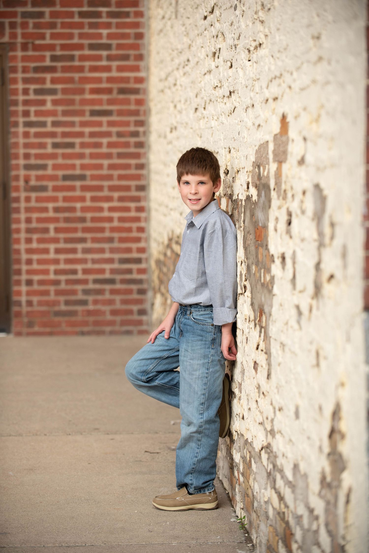 Child in denim and gray shirt leans playfully against distressed white wall near brick surface.