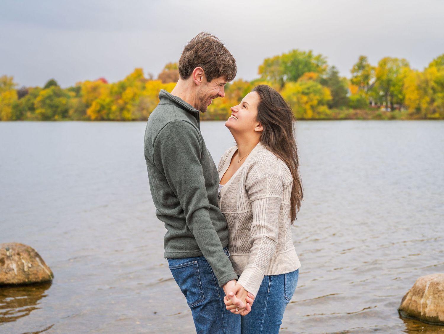 A couple shares a romantic moment by a lake during autumn with fall foliage in the background.