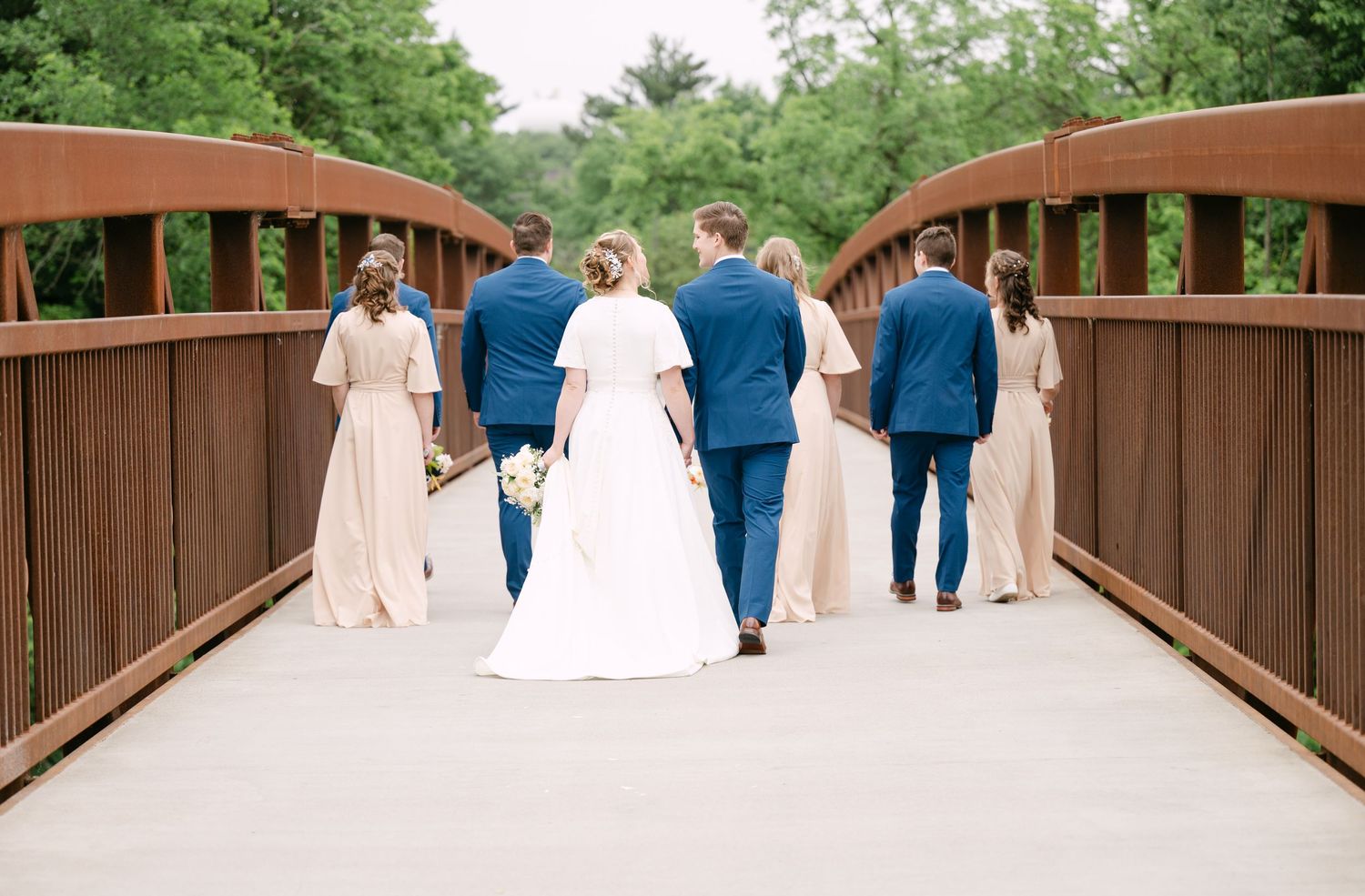 Wedding party walking across rustic wooden bridge surrounded by lush greenery.