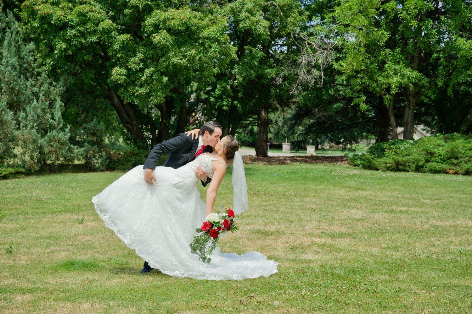 Groom dips bride for romantic kiss outdoors on green lawn surrounded by lush trees.