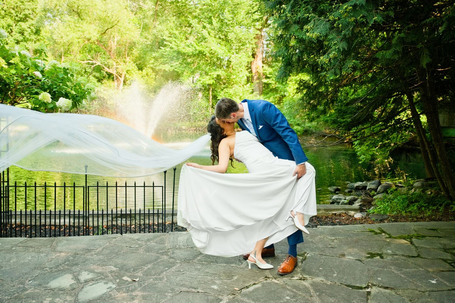 Groom dips bride in a romantic kiss outdoors, her veil flowing gracefully, surrounded by lush green trees.