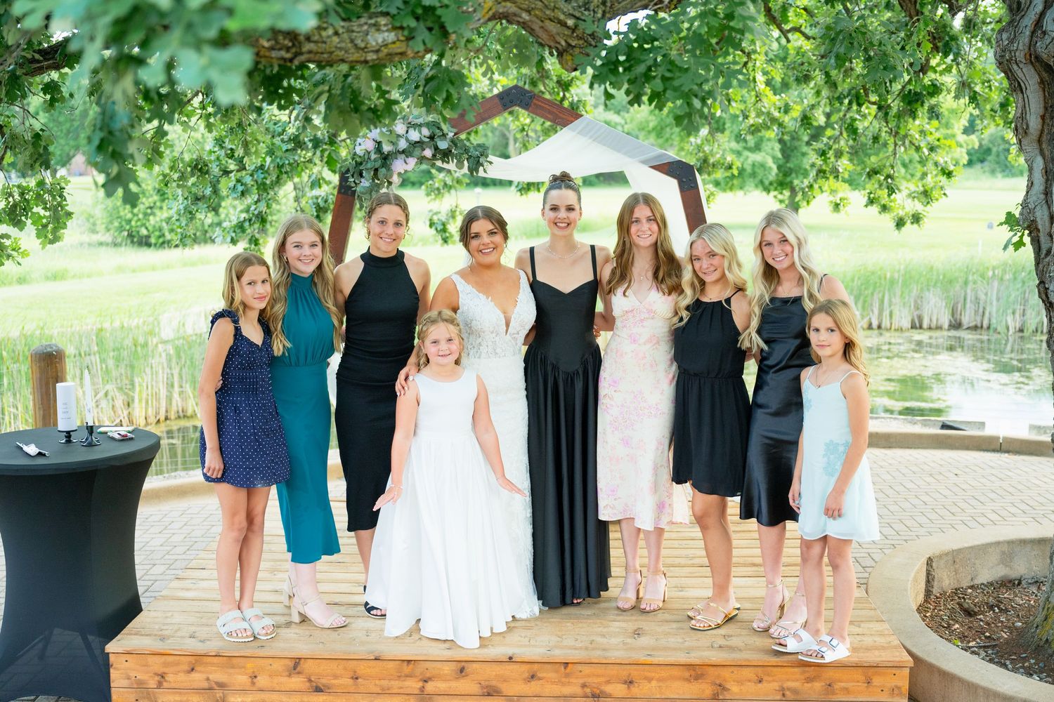 Bride in white gown posing with bridesmaids and flower girls outdoors at wedding reception.