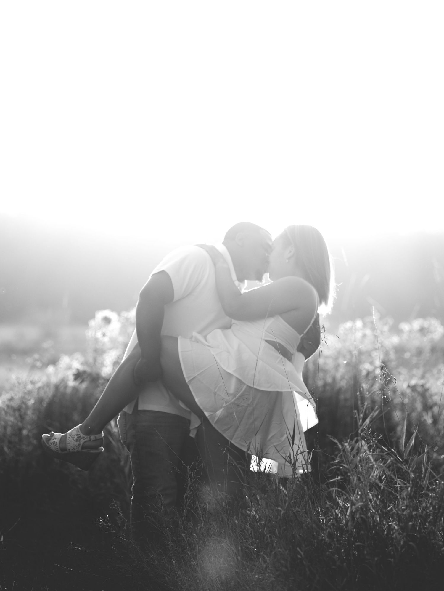 Central Minnesota Best Engagement Photographer - Black and white photo of a couple kissing in a sunlit field, surrounded by tall grass.