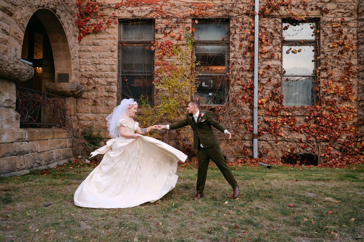 Central MN Wedding Photos | Bride in vintage gown twirls with groom outside stone building with autumn ivy.