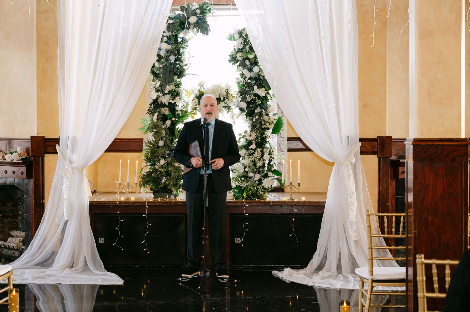 Central MN Wedding Photos | Officiant stands at floral arch altar with white drapes at elegant wedding venue.