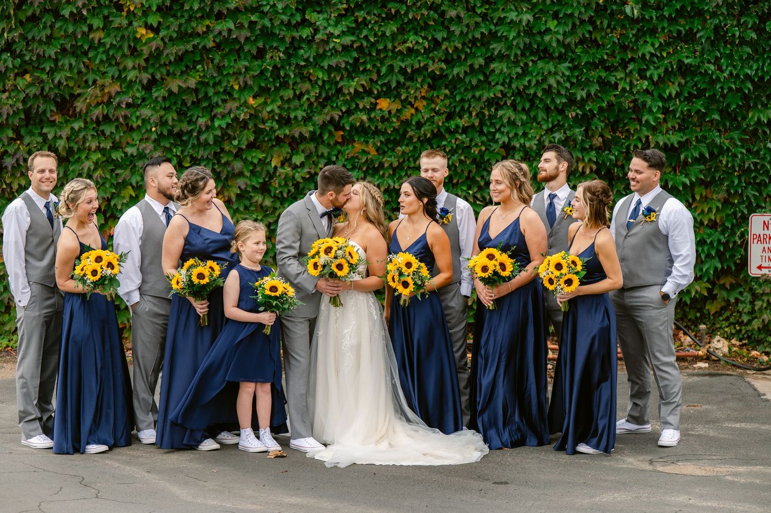 Central MN Wedding Photos | Wedding party poses together with bridesmaids in navy dresses holding sunflower bouquets against an ivy wall.