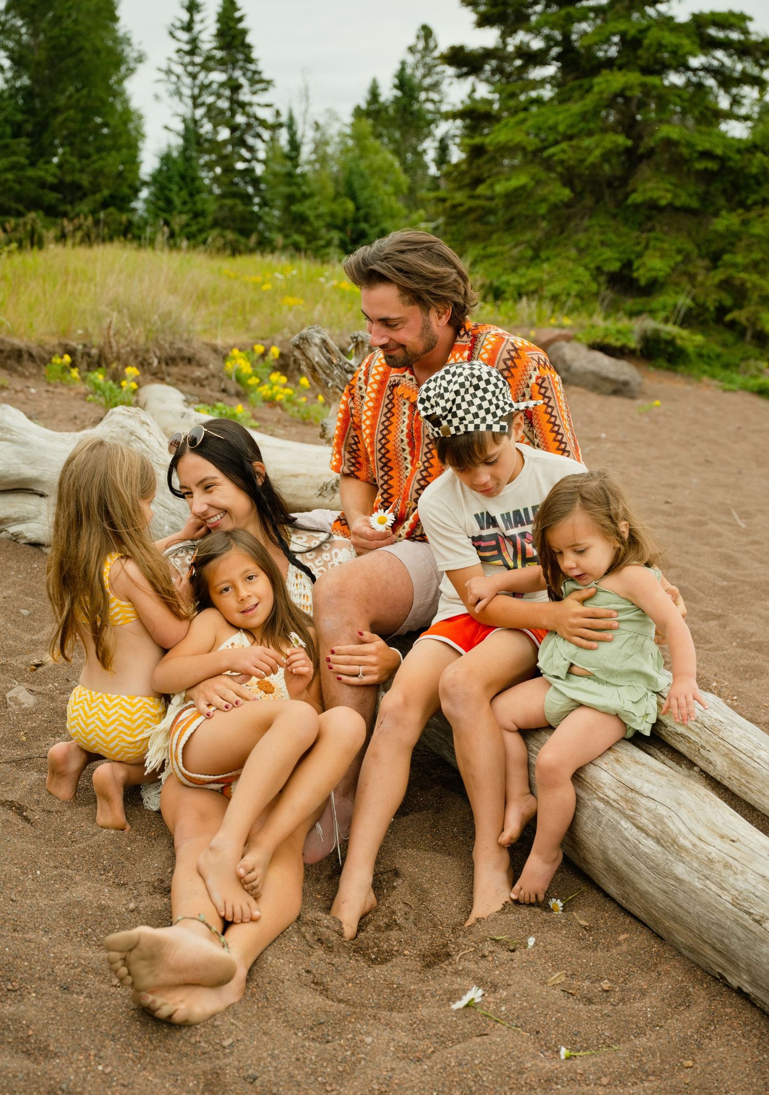 A joyful family with young children sitting together on a sandy beach log surrounded by pine trees. Grand Marais Family Photography