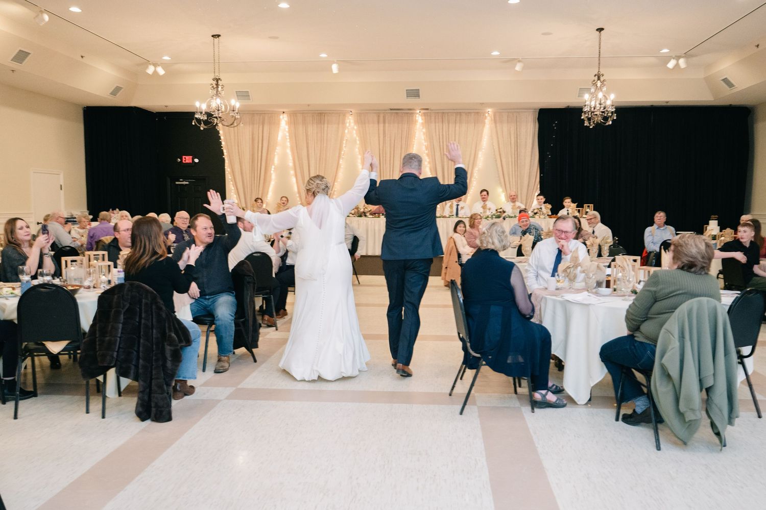 Bride and groom enter wedding reception hall with raised hands as guests applaud at elegantly decorated venue. Northern Lights Milaca MN