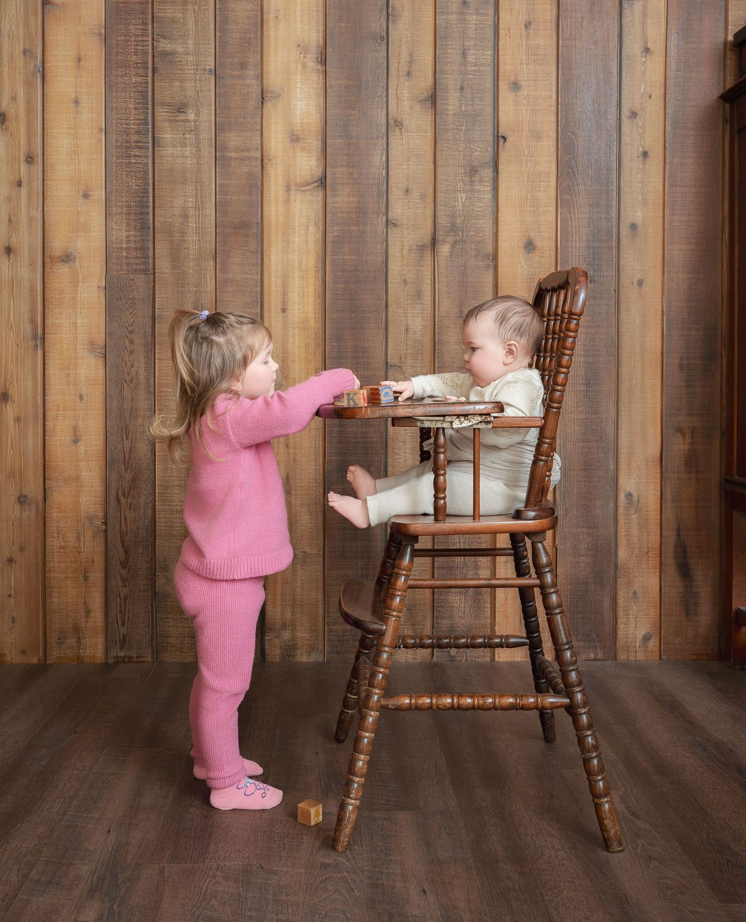 Two toddlers interact near a wooden high chair against a rustic wood panel wall, one in pink feeding the other.