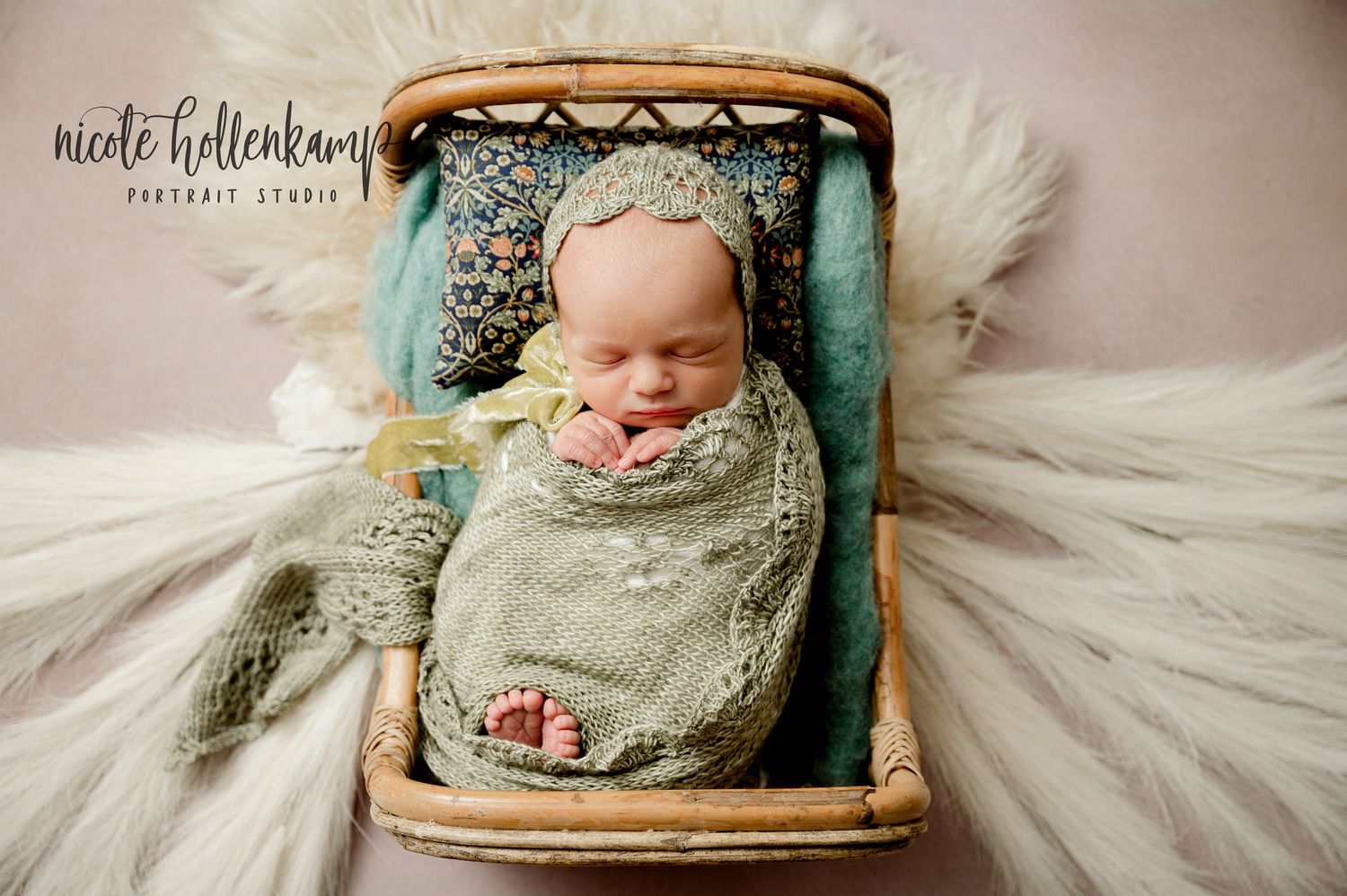 Newborn swaddled in sage green wrap sleeps peacefully in vintage wooden basket surrounded by soft white fabric.
