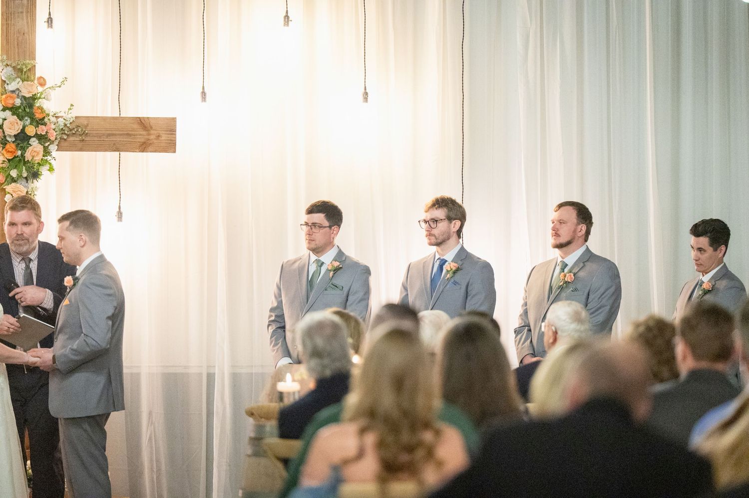 Groomsmen in gray suits stand at decorated altar with floral arrangements during indoor wedding ceremony.